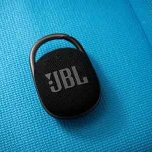 Black JBL portable wireless speaker with carabiner on textured blue background. Ideal for travel and outdoor use.